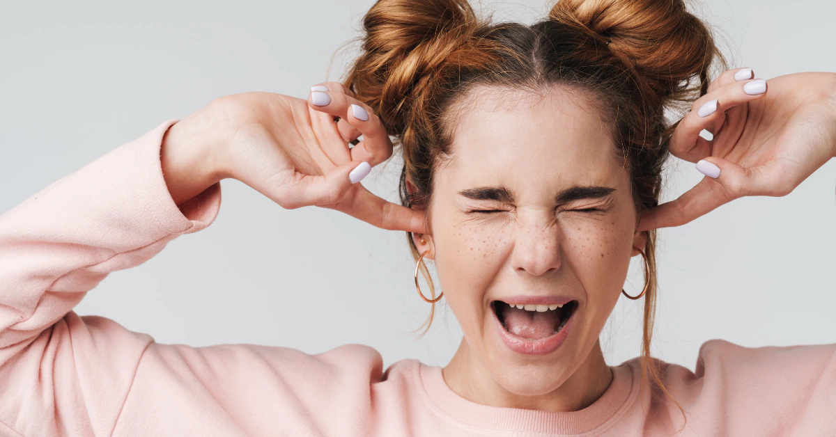 Vrouw in roze trui die haar oren dicht drukt en haar ogen dichtknijpt als reactie op hard geluid.