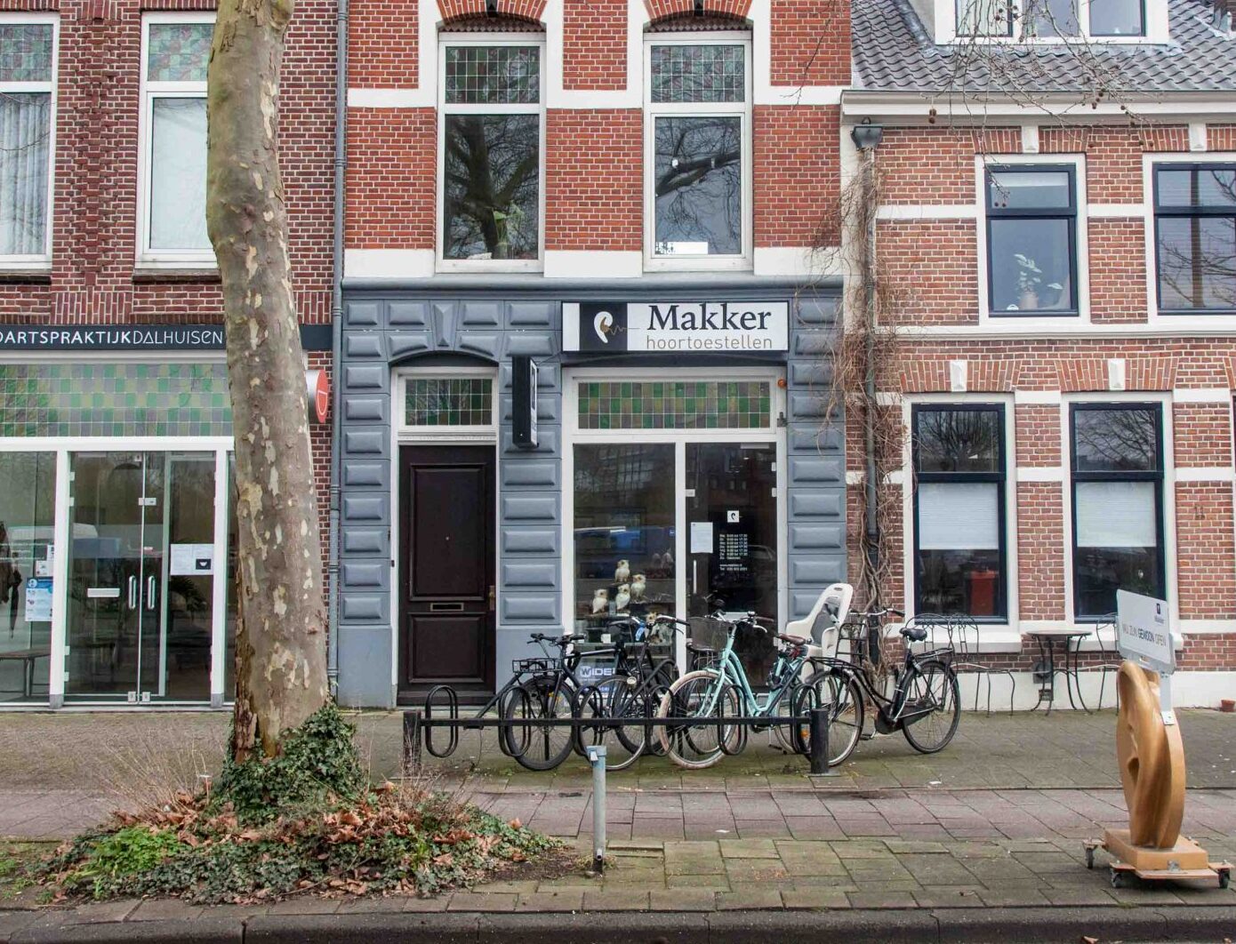 Vooraanzicht van de winkel "Makker hoortoestellen" met fietsenrekken en geparkeerde fietsen op de stoep.
