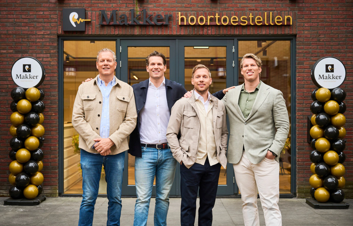 Vier mannen poseren voor de ingang van Makker hoortoestellen, geflankeerd door ballonnen.