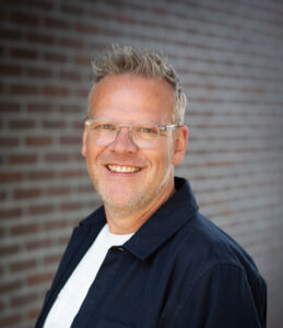 Man met bril en donker jasje lacht naar de camera voor een muur van rode bakstenen.