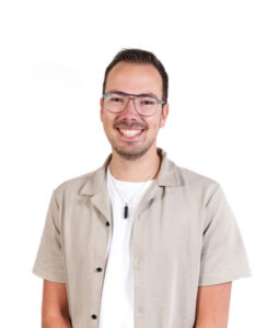 Man met bril en beige overhemd poseert glimlachend voor een witte achtergrond.