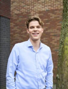 Jonge man in lichtblauw overhemd lacht terwijl hij buiten voor een bakstenen muur poseert.