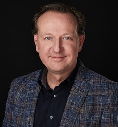 Portret van Franck van Popta in een blauw geruit colbert tegen een zwarte achtergrond.