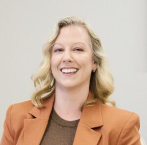 Vrouw met blond, krullend haar en een oranjebruine blazer lacht naar de camera tegen een neutrale achtergrond.