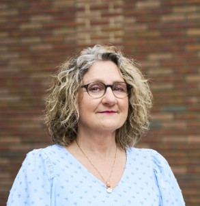Vrouw met bril en krullend haar poseert voor een muur van rode bakstenen.