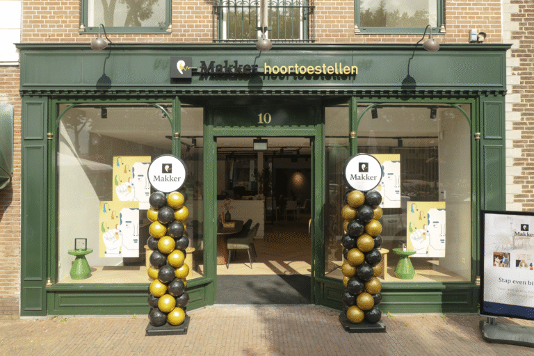Gevel van Makker hoortoestellen met groene kozijnen, ballonnenzuilen en open ingang.