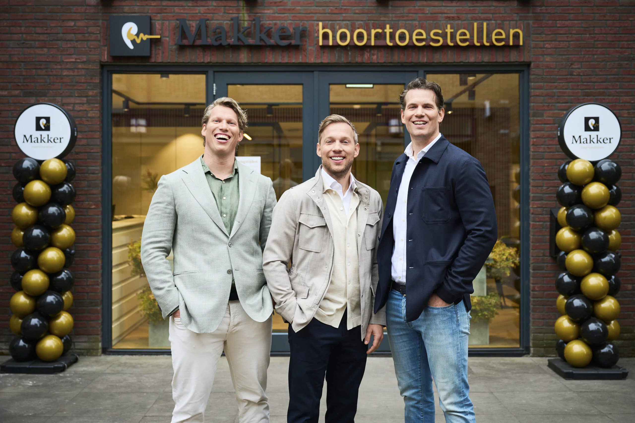 Drie lachende mannen poseren voor de ingang van Makker hoortoestellen, met feestelijke ballondecoraties naast de deur.