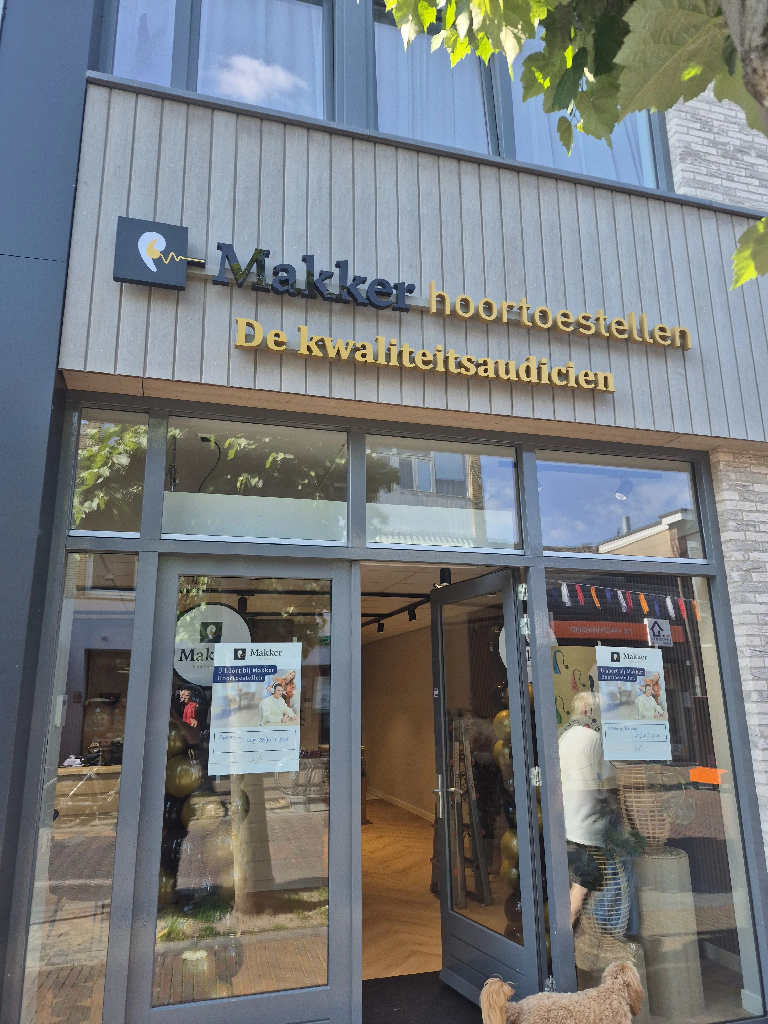 De gevel van de winkel "Makker hoortoestellen", een kwaliteitsaudicien, met geopende deur en raamstickers.