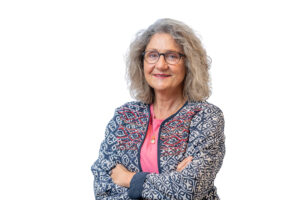 Een vrouw met grijs krullend haar, gekleed in een kleurrijke jas, staat met gekruiste armen en glimlacht naar de camera.