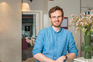 Een persoon in een blauwe blouse glimlacht terwijl hij naast een vaas met bloemen in een moderne en lichte kantoorruimte staat.