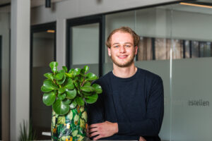 Een jonge man glimlacht naast een groene kamerplant in een moderne kantooromgeving.