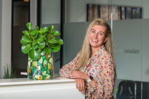 Een lachende vrouw in een bloemetjesblouse leunt tegen een wit bureau met een groene plant in een kleurrijke pot erop, in een moderne kantooromgeving.