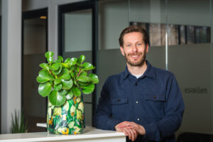 Een man met een blauwe blouse glimlacht naar de camera terwijl hij naast een groene plant in een kleurrijke pot staat.