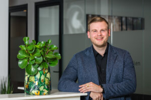 Man in een blauw jasje leunt glimlachend op een tafel naast een kleurrijke kamerplant in een modern kantoor.