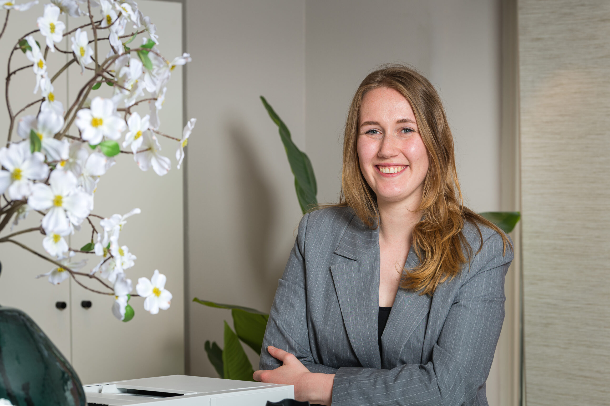 Een lachende vrouw in een grijs pak staat bij een bureau met witte bloemen op de voorgrond.