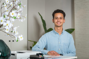 Een glimlachende jongeman zit achter een balie met een bloemenvaas en een pinapparaat.