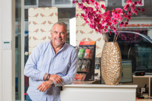 Een man leunt glimlachend op een balie in een winkel, naast een vaas met roze bloemen en een display van Noizezz producten.