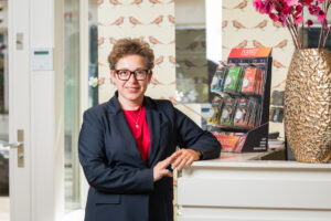 Vrouw in een zakelijk pak poseert naast een display van Noizezz oordopjes op een receptiebalie, met een gouden vaas en bloemen op de achtergrond.