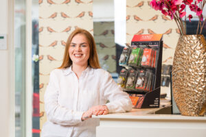 Een vrouw in een witte blouse glimlacht naar de camera naast een display met Notezz oordopjes in een winkel met vogelbehang.