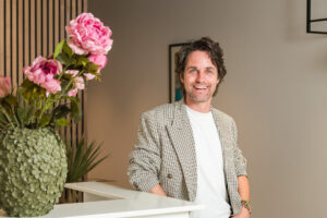 Een glimlachende man in een geruite blazer staat naast een vaas met grote roze bloemen in een stijlvolle, moderne ruimte.