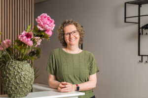 Een lachende vrouw met krullend haar en een groene top staat naast een vaas met roze bloemen.