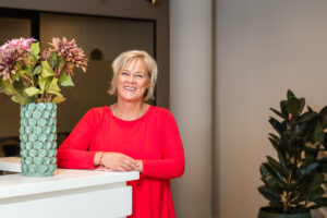 Vrouw in rode blouse lacht terwijl ze leunt op een wit bureau naast een vaas met bloemen in een moderne kantooromgeving.