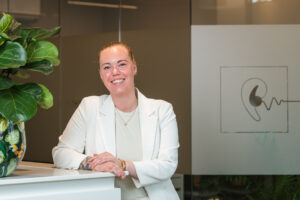 Een glimlachende vrouw in een witte outfit poseert naast een balie met een plant in een moderne kantooromgeving.