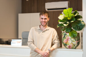 Een lachende jonge man leunt tegen een balie naast een decoratieve potplant in een modern kantoorinterieur.