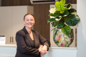 Een lachende vrouw in een donkere blazer leunt op een balie naast een kleurrijke potplant in een moderne kantooromgeving.