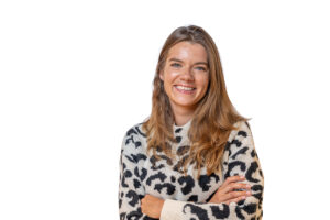 Portret van een lachende vrouw met lang haar in een trui met luipaardmotief, staand voor een witte achtergrond.
